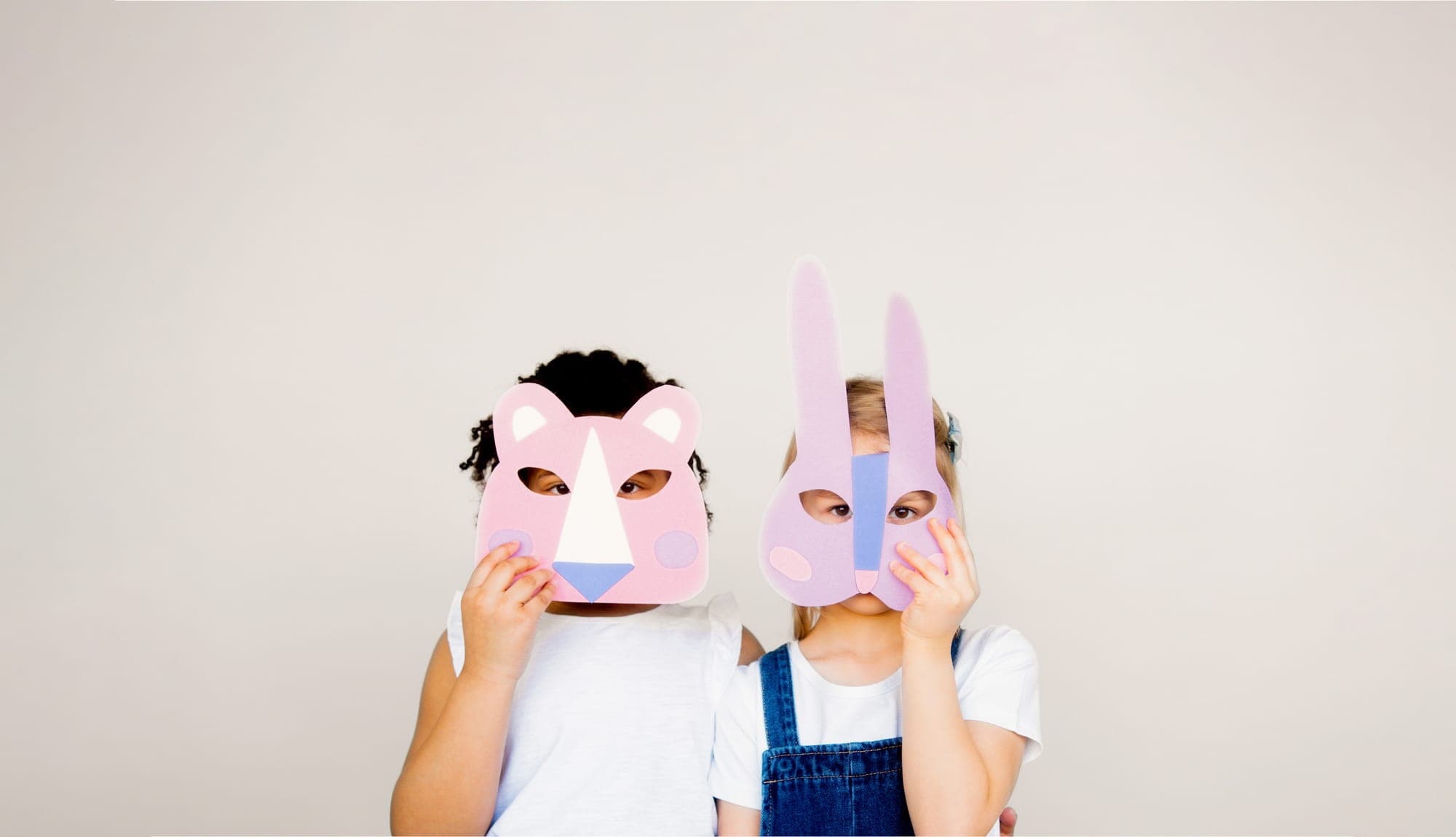 Dos niñas juegan con máscaras de papel de un oso y un conejo frente a una pared clara.